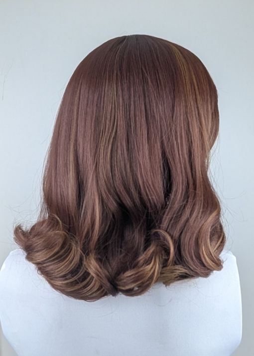 A warm brown wig with golden blonde highlights throughout. Styled into a long bob thats sleek from the roots, ending in large curls. The full blunt fringe is sleek.