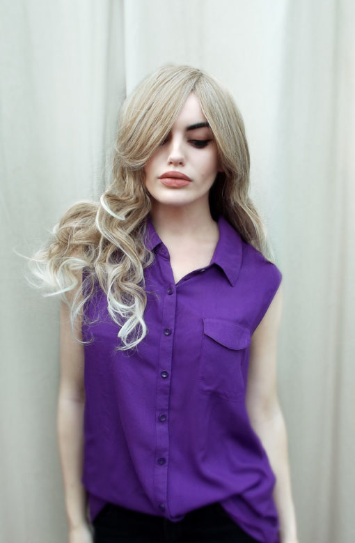 Blonde long curly wig with bangs. Natural blonde is simple and elegant. Dark blond roots that blend into a golden blonde shade. The ends of the hair have a sun-kissed effect. The fringe is long and thick to add extra layers. Sleek from the roots and becomes big curls that fall just past the shoulders.