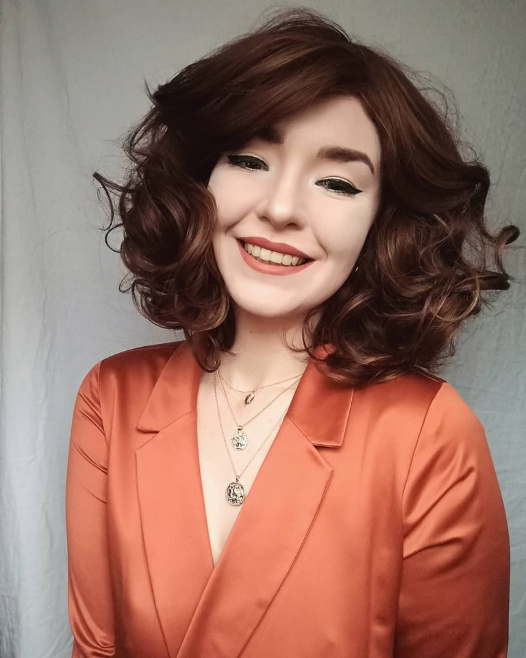 A warm brown wig with golden blonde highlights throughout. Styled into a long bob thats sleek from the roots, ending in large curls. The full blunt fringe is sleek.
