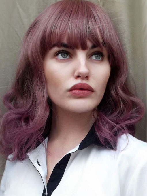 Lilac curly bob wig with bangs. Berry jam is a long bob cut with a natural twist of caramel hues from the roots, that blends into a sweet and sugary mixture of dusky lilacs and rose tones. The ends have a dip-dye of dark lilac. Set in loose curls to give it plenty of movement. Falling to the shoulders with a long, full fringe.