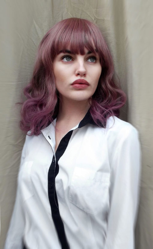 Lilac curly bob wig with bangs. Berry jam is a long bob cut with a natural twist of caramel hues from the roots, that blends into a sweet and sugary mixture of dusky lilacs and rose tones. The ends have a dip-dye of dark lilac. Set in loose curls to give it plenty of movement. Falling to the shoulders with a long, full fringe.
