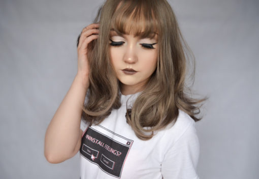Grey curly wig with bangs. Sepia is silky, soft, and creates an effect of subtle fullness. A mixture of warm brown and ash blonde, sleek with curls at the ends to generate texture and movement.