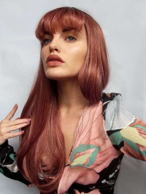 Red long straight wig with bangs. Chantilly is pretty and alluring. Sleek with red and pink golden tones, that nods to rose gold hues. With dark brown shadowed roots that gives a grown out natural finish.
