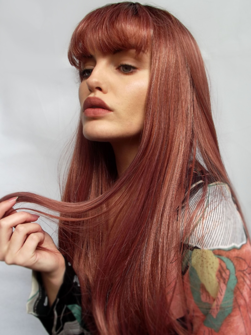 Red long straight wig with bangs. Chantilly is pretty and alluring. Sleek with red and pink golden tones, that nods to rose gold hues. With dark brown shadowed roots that gives a grown out natural finish.