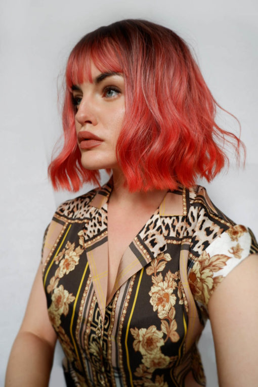 Coral wavy bob wig with bangs. Bellini is a light choppy bob. Brown shadowed roots give a natural feel to the look. The coral colour is complemented by its dropped out waves.