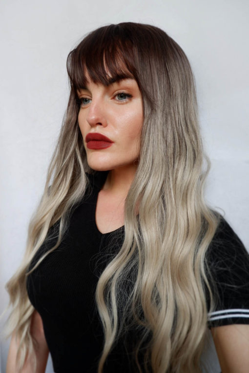 Blonde wavy long wig with bangs. Haunt comes to life with brown grown out roots. They blend into a dirty blonde tone, with a sandy shade as dip dye ends. The style has a soft wave with a more defined braid wave through the length.