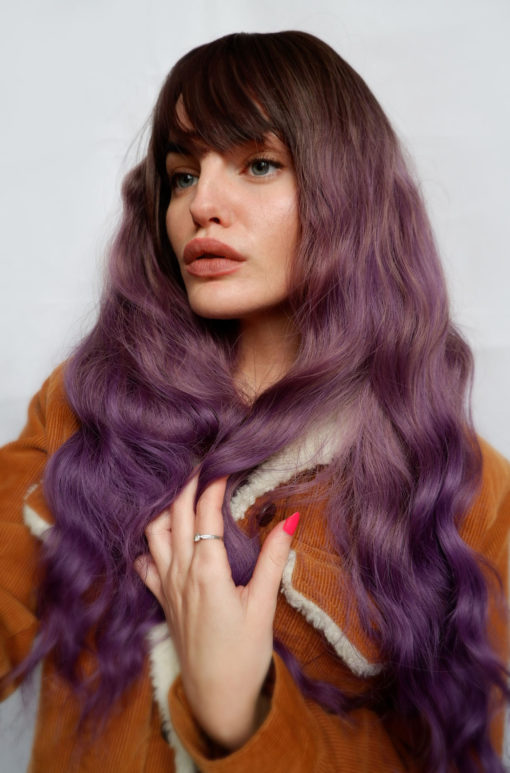 Purple long wavy wig with bangs. Calliope takes shades of purple in different directions. Sharp and uneven, it creates a cool, edgy look. Warm brown shadowed roots blend into a washed-out purple ombre. The ends have been dip dyed in a rich deep purple, the face is famed with a fringe and soft loose long waves that fall to the waist.