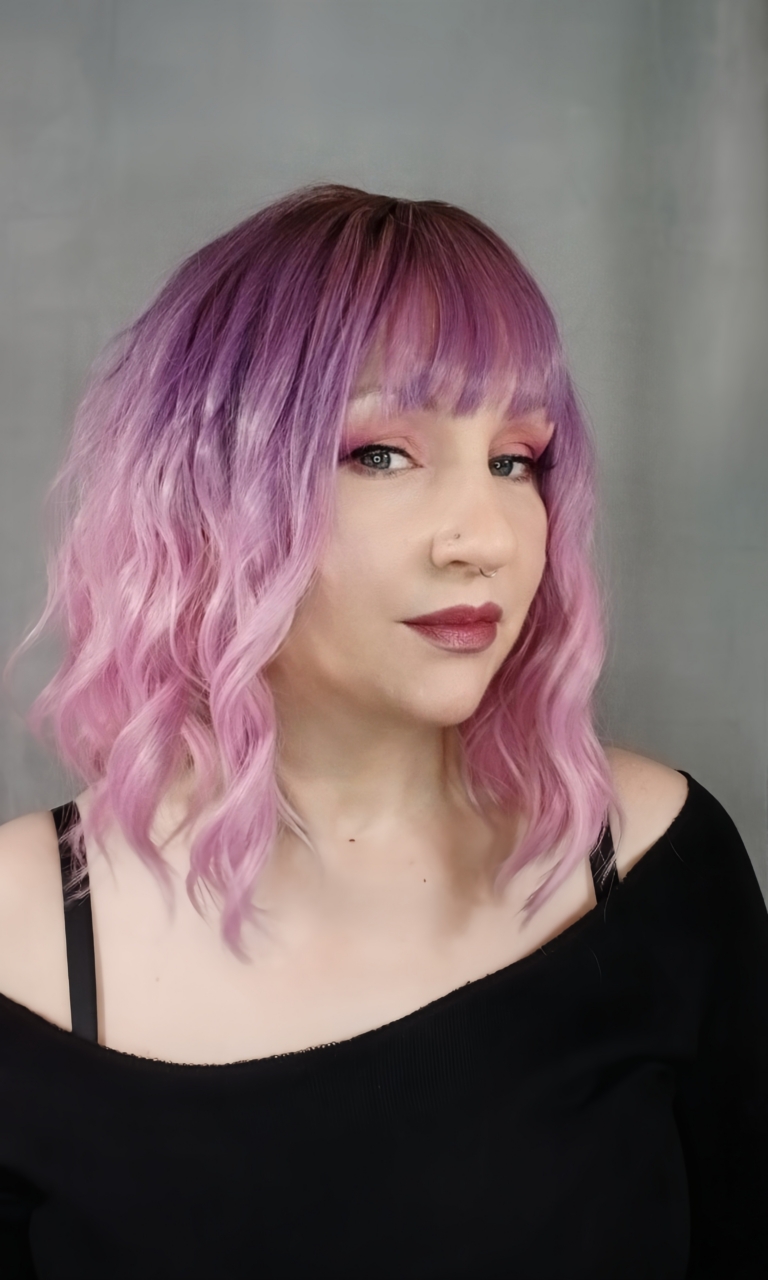 This wig has brown shadowed roots that melt into a dark lilac colour into a soft pink ombre. A bob styled into loose waves with a sleek blunt fringe.
