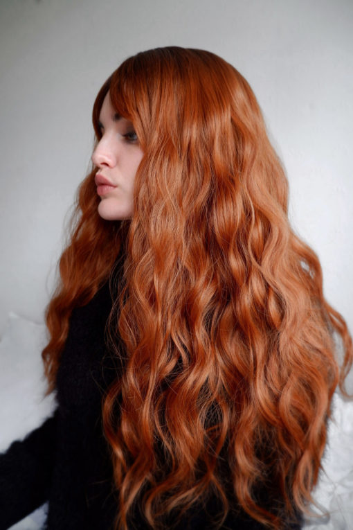 Burnt orange long wavy wig with bangs. Hinode is a spiciness delight. Dark brown shadowed roots blend into a mix of burnt orange and auburn. Long loose waves echos styles from the 70s.