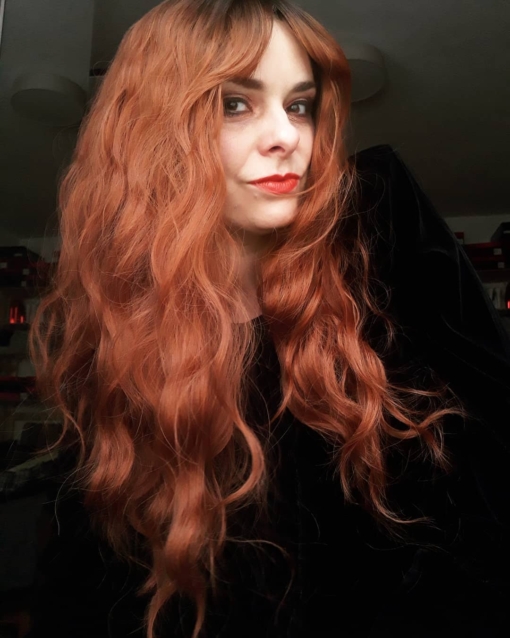 Burnt orange long wavy wig with bangs. Hinode is a spiciness delight. Dark brown shadowed roots blend into a mix of burnt orange and auburn. Long loose waves echos styles from the 70s.