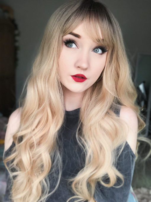 Long blonde curly wig with bangs. Inspired from looks of the 60s and 70s era. Eliza is natural with brown shadowed roots that melt into a sun-kissed blonde. Long beachy waves and curtain bangs take us back in time.