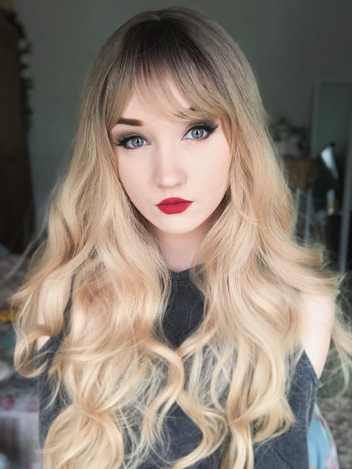 Long blonde curly wig with bangs. Inspired from looks of the 60s and 70s era. Eliza is natural with brown shadowed roots that melt into a sun-kissed blonde. Long beachy waves and curtain bangs take us back in time.