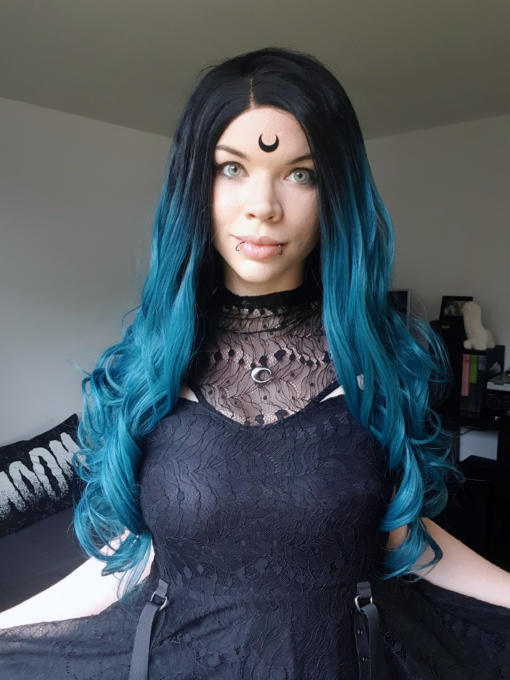 Long curly blue teal lace front wig. This eye-catching blueish/green concoction of colours is Apatite. Black roots add a natural edge to the style.