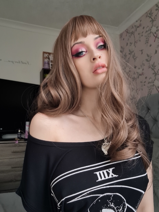 Dark blonde long curly wig with bangs. Jana is a natural long style with very loose tumbling curls. The colours are a mix of dark blonde and very subtle muted pinks. From roots to tips. A sleek fringe frames the face.