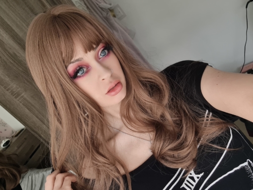 Dark blonde long curly wig with bangs. Jana is a natural long style with very loose tumbling curls. The colours are a mix of dark blonde and very subtle muted pinks. From roots to tips. A sleek fringe frames the face.