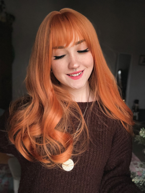 Ginger wavy long wig with bangs. Sunbeam is an unusual orange colour blend of golden copper from roots to tips. A sleek style with gentle curls at the ends with a light fringe, produces this eye-catching look.