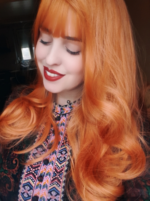 Ginger wavy long wig with bangs. Sunbeam is an unusual orange colour blend of golden copper from roots to tips. A sleek style with gentle curls at the ends with a light fringe, produces this eye-catching look.
