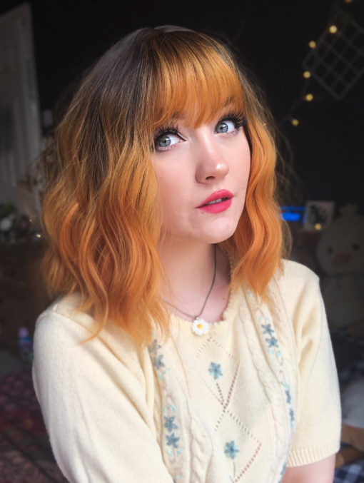 Orange wavy bob wig with bangs. Amber is a low maintenance choppy bob. Warm sunset tones with brown shadowed roots give a natural feel to the look. Beachy waves give texture to the style with an airy fringe.