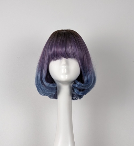 Lilac and blue curly bob with bangs. Carina combines a sleek dark lilac colour, that melts into a denim blue that curls under. Light brown shadow roots give a natural grown out feel, with a wispy fringe.