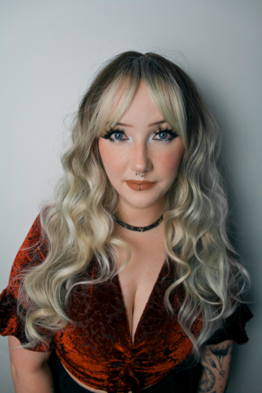 A mix of dark brown and blonde sleek roots that blend into a blonde and hazel colour. The fringe is long and sleek that can be parted for curtain bangs. It is styled in big loose curls.