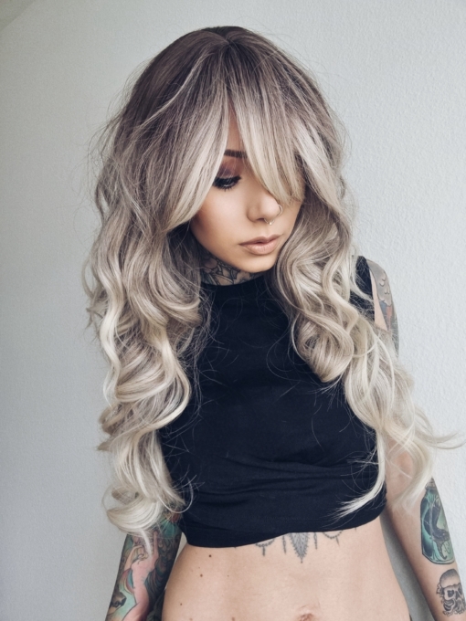 A mix of dark brown and blonde sleek roots that blend into a blonde and hazel colour. The fringe is long and sleek that can be parted for curtain bangs. It is styled in big loose curls.