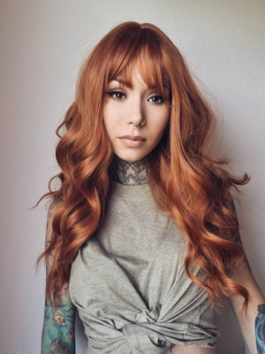 This wig has brown shadow roots that blend into a natural muted auburn colour. Styled in loose barrel curls falling to the waist. The fringe is sleek and blunt.