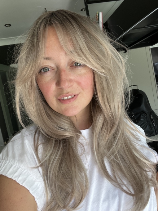 This natural wig comes with brown shadowed roots that blend into a ash blonde with a hint of rose gold. Its styled into very loose curls. With a long thick sleek fringe that can be worn as curtain bangs.