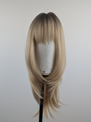 A long and sleek wig with natural dark brown shadowed roots. They blend into a honey blond with sterling blonde highlights. Styled into sleek long layers and a long fringe that can swept to the side or parted into curtain bangs.