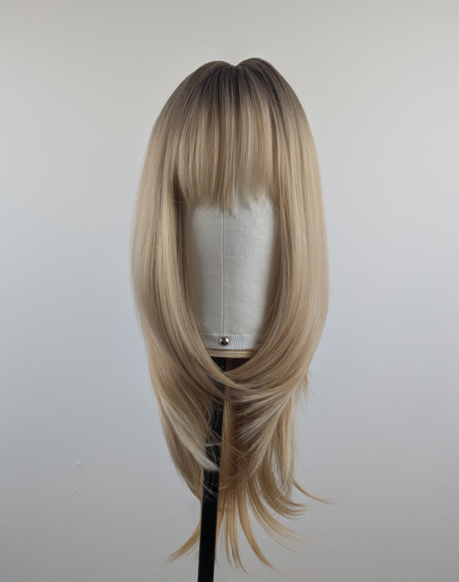 A long and sleek wig with natural dark brown shadowed roots. They blend into a honey blond with sterling blonde highlights. Styled into sleek long layers and a long fringe that can swept to the side or parted into curtain bangs.