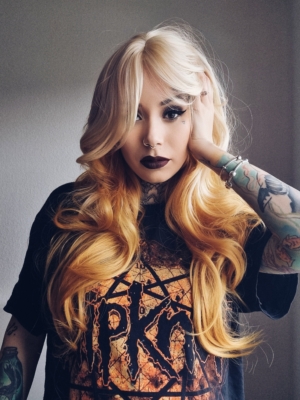 A light blonde colour from crown of the head and the fringe. Underneath is a soft natural ginger sunshine hue. Styled sleek and long into waves throughout the lengths of the hair with cut in layers. A blunt fringe frames the face.