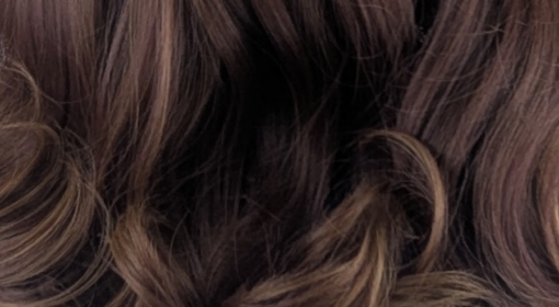 A warm brown wig with golden blonde highlights throughout. Styled into a long bob thats sleek from the roots, ending in large curls. The full blunt fringe is sleek.